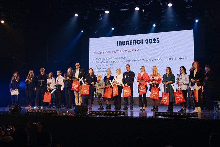 Na zdjęciu widzimy grupę kilkunastu osób stojących obok siebie na scenie. W tle znajduje się duży biały ekran z czarnym napisem Laureaci 2025, poniżej wymienione imiona i nazwisk. Większość z nich trzyma w dłoniach pomarańczowe torby z białym napisem „Moje zakupy niosą pomoc” oraz czerwone róże. Dwie z nich dodatkowo trzymają puchar laureata. Uczestnicy są zwróceni przodem do widza i pozują do wspólnego zdjęcia.