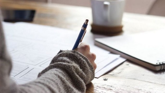 - Na pierwszym planie ręka, w której dłoni znajduje się długopis. Na biurku leżą dokumenty, otwarty notes, stoi tam również filiżanka.
