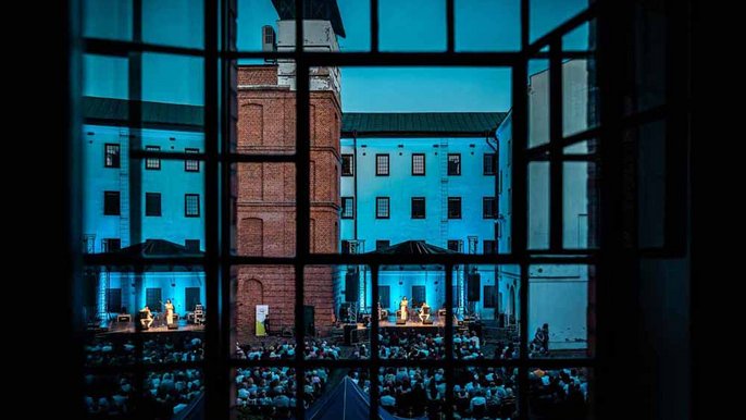  - Na pierwszy planie stare, fabryczne okno przed które widać dziedziniec Centralnego Muzeum Włókiennictwa w Łodzi. Na dziedzińcu duża grupa siedzących ludzi zwróconych w stronę sceny, na której grają muzycy.