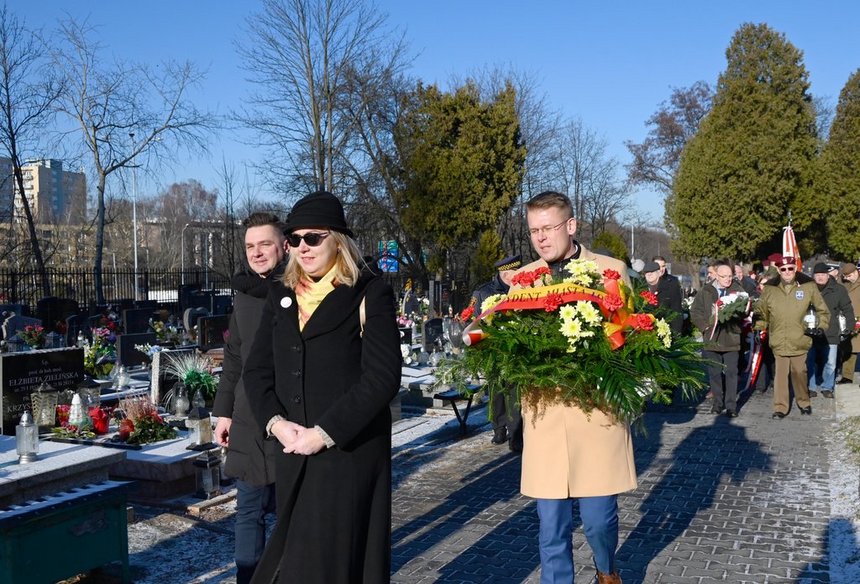 Alejka na cmentarzu. Kondukt ludzi niosących wiązanki. Na pierwszym planie wiceprezydent Łodzi Adam Wieczorek niosący wieniec z wstęgą na napisem Prezydent Miasta Łodzi. Obok niego idzie Przewodniczący Rady Miejskiej w Łodzi Marcin Gołaszewski.