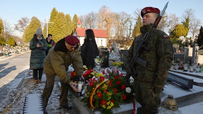  - Cmentarz. Przy grobie weteranów powstania Wielkopolskiego stoi warta honorowa. Na pierwszym planie żołnierz na baczność z karabinem i bagnetem. Na grobie kwiaty. Drugi żołnierz stawia na grobie zapalony znicz. W tle widzać tablicę grobową na napisem Rodzina Walczaków.