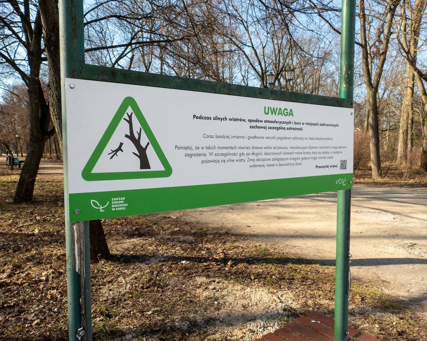 Tablica informująca o zagrożeniu przy wejściu do parku. Na tablicy grafika trójkąt z zielona obwódką, w środku fragment drzewa z odłamującą się gałęzią. Napis Uwaga, podczas silnych wiatrów zachowaj szczególną ostrożność.