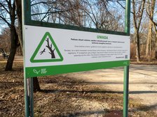 Tablica informująca o zagrożeniu przy wejściu do parku. Na tablicy grafika trójkąt z zielona obwódką, w środku fragment drzewa z odłamującą się gałęzią. Napis Uwaga, podczas silnych wiatrów zachowaj szczególną ostrożność.