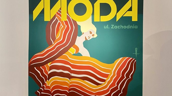 - W sali wystawowej wisi wielkoformatowa reprodukcja, a na niej wizerunek kobiet w kolorej sukni. Na górze obrazu napis "MODA".