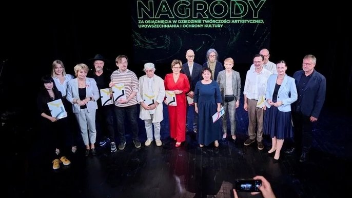- Na scenie teatralnej stoi grupa ludzi, którzy w rękach trzymają dyplomy. Są to laureaci nagród w 2023 roku. W tle napis: Nagrody za osiągnięcia w dziedzinie twórczości artystycznej, upowszechnia i ochrony kultury.