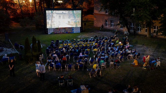 - Kadr wypełniają leżaki z widzami na łące. Osoby oglądają film wyświetlany na dużym ekranie. Wieczór jest wczesny. Mrok jeszcze nie zapadł.