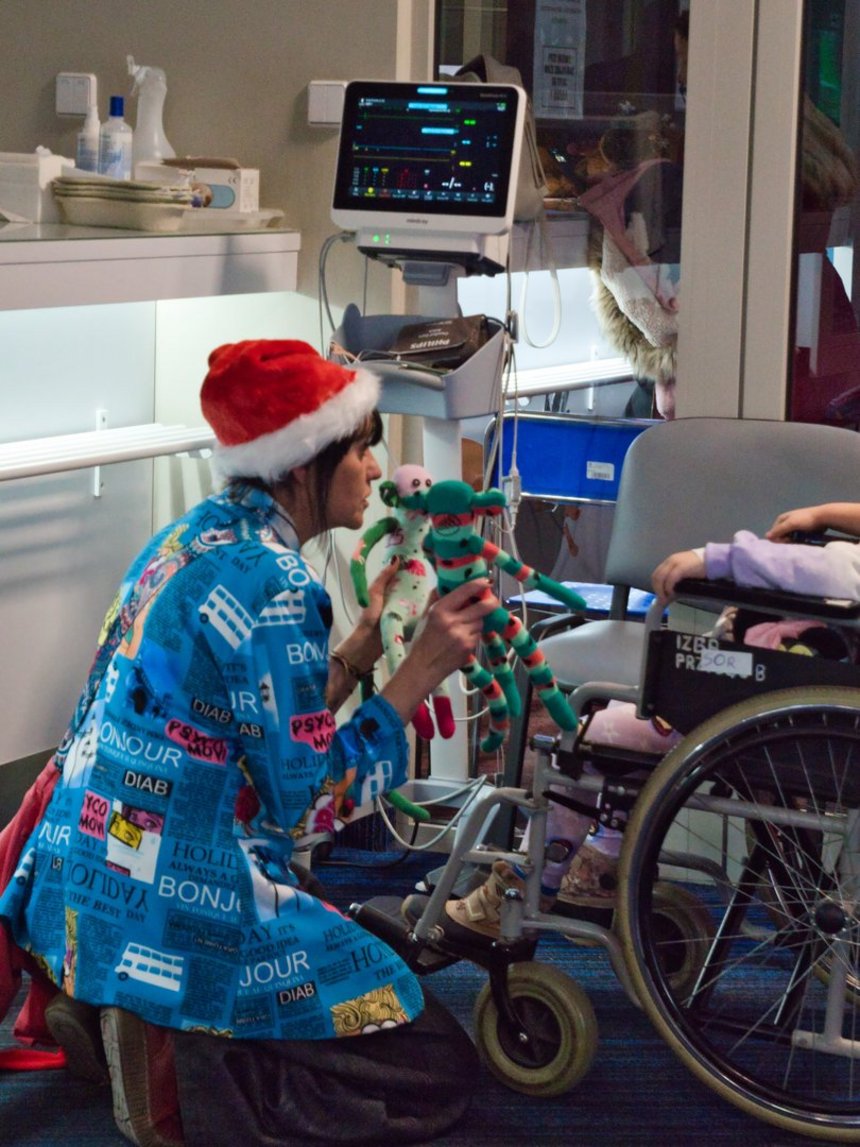 Na zdjęciu widzimy dorosłą osobę w czapce Mikołaja, kucającą przy dziecku na wózku inwalidzkim, w rękach trzyma i pokazuje dziecku dwie kolorowe materiałowe maskotki w kształcie małpek. W tle widać sprzęt medyczny i monitor.