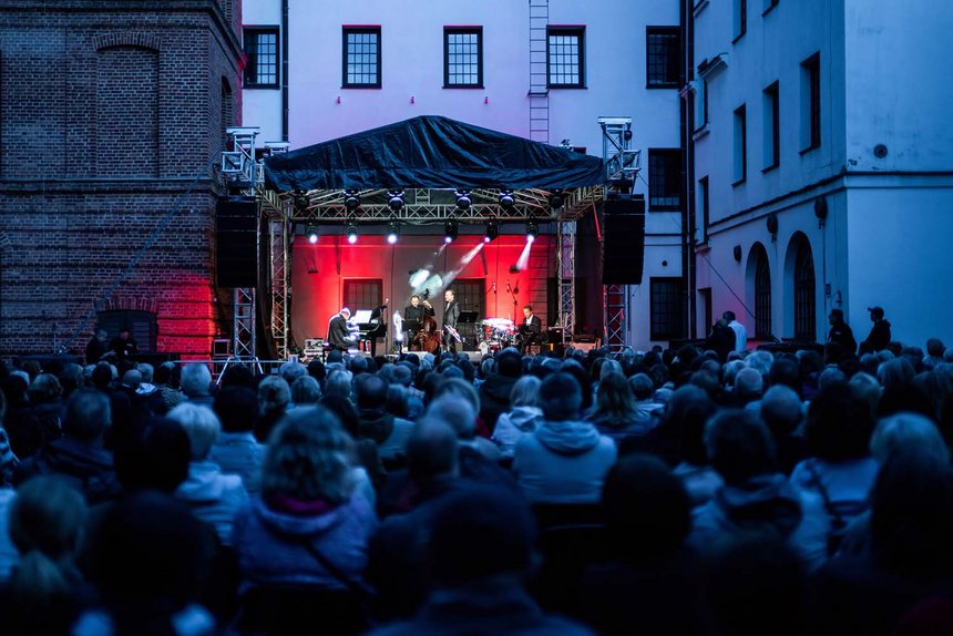 W centralnym punkcie scena plenerowa oświetlona jasnym, czerwonym i białym światłem, na niej muzycy. Na pierwszym planie widoczna publiczność zwrócona w stronę sceny.