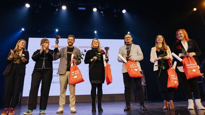  - Na zdjęciu widać siedem osób stojących obok siebie na scenie. W tle znajduje się duży biały ekran. Pięć osób trzyma w dłoniach pomarańczowe torby z białym napisem „Moje zakupy niosą pomoc” oraz czerwone róże. Trzy z nich dodatkowo puchar laureata. Uczestnicy są zwróceni przodem do widza i pozują do wspólnego zdjęcia.