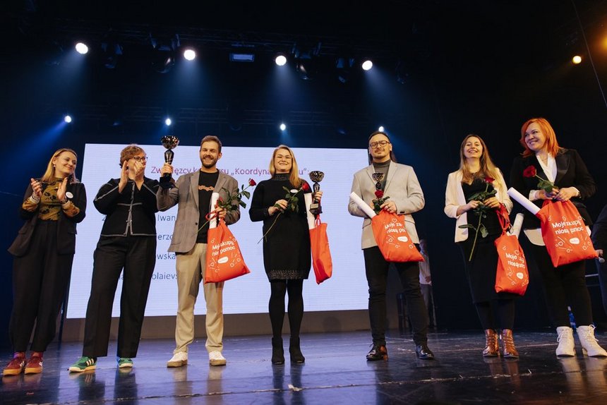 Na zdjęciu widać siedem osób stojących obok siebie na scenie. W tle znajduje się duży biały ekran. Pięć osób trzyma w dłoniach pomarańczowe torby z białym napisem „Moje zakupy niosą pomoc” oraz czerwone róże. Trzy z nich dodatkowo puchar laureata. Uczestnicy są zwróceni przodem do widza i pozują do wspólnego zdjęcia.