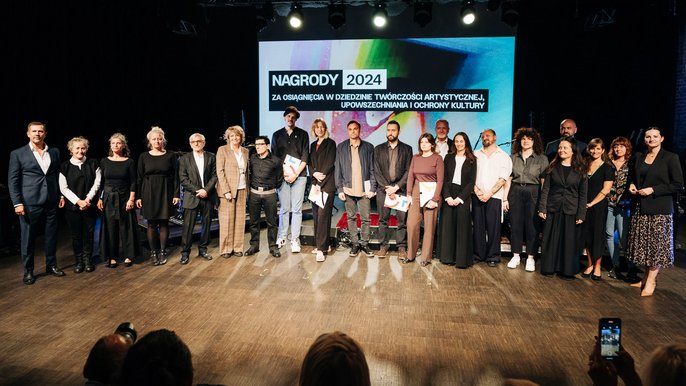 - Na zdjęciu grupa osób, stojąca na scenie. Laureaci nagrody stoją w rzędzie na wprost. Ponad nimi napis: Nagrody 2024 za osiągnięcia w dziedzinie twórczości artystycznej, upowszechniania i ochrony kultury.