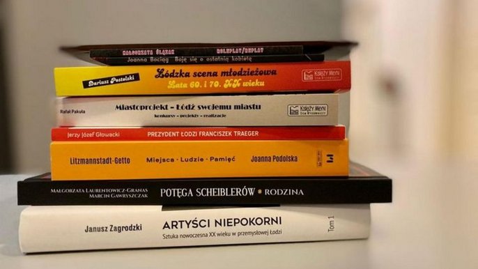 - W bliskim planie stos książek, na których grzbietach widać informacje o tytułach i autorach.