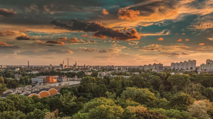 - Panorama Łodzi na tle zachodzącego słońca. Ponad parkiem wysokie zabudowania na linii horyzontu.