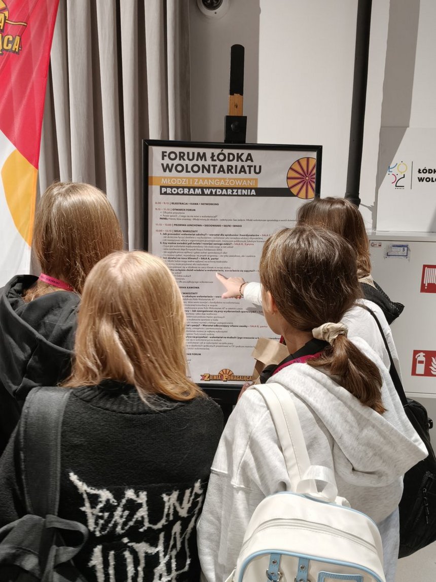 Na zdjęciu widzimy grupę młodzieży wskazującą plakat, który znajduje się przed nimi na staludze. Na plakacie wielkimi literami napisane jest: Forum Łódka Wolontariatu. Młodzi i Zaangarzowani. Program Wydarzenia. 