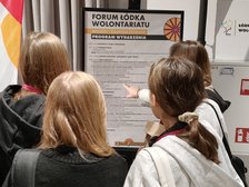 Na zdjęciu widzimy grupę młodzieży wskazującą plakat, który znajduje się przed nimi na staludze. Na plakacie wielkimi literami napisane jest: Forum Łódka Wolontariatu. Młodzi i Zaangarzowani. Program Wydarzenia. 